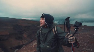 Yağmurluklu adam, volkanda yürüyen İskandinav için sopaları tutuyor, mesafeye bakıyor.