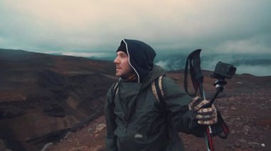 Yağmurluklu adam, volkanda yürüyen İskandinav için sopaları tutuyor, mesafeye bakıyor.