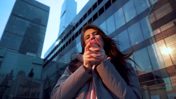 Girl drinks coffee among glass skyscrapers at sunset in windy weather. — Αρχείο Βίντεο