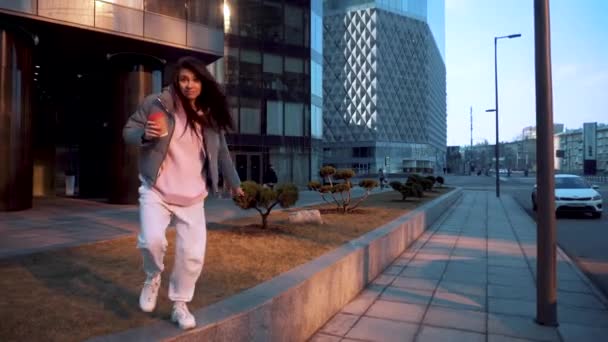 A girl in a tracksuit with coffee dances in a big city among the glass buildings — Stockvideo