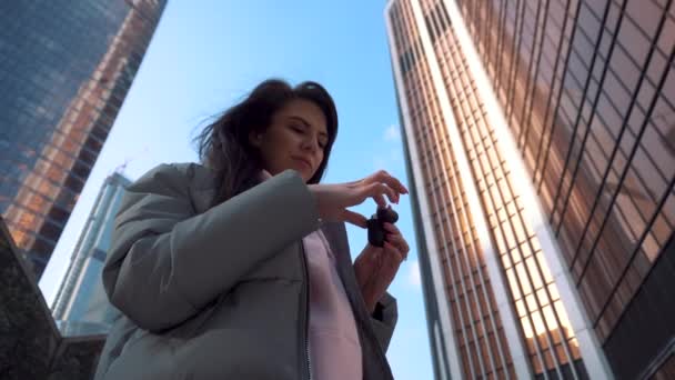 Girl pulls out and puts on wireless headphones among the skyscrapers — Αρχείο Βίντεο