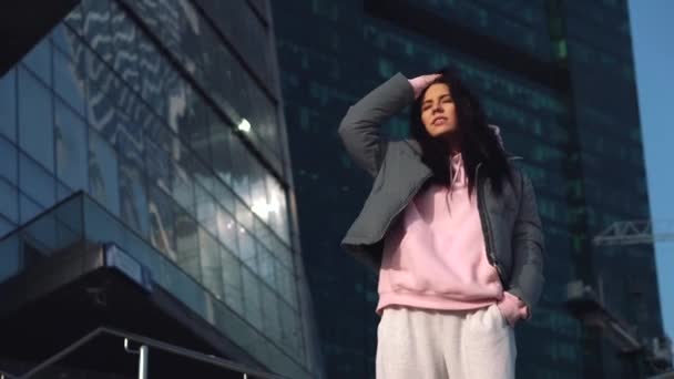 Young girl on the background of glass buildings straightens her hair in windy weather — Αρχείο Βίντεο