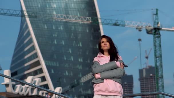 한 소녀는 고층 건물, 기중기 및 산책로 가 있는 현대 건축 현장의 배경에서 있다 — 비디오