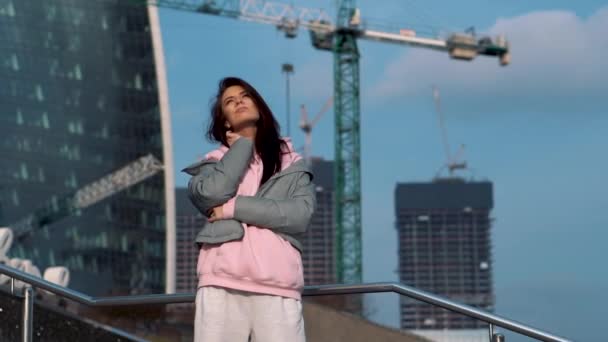 A girl stands against the backdrop of a modern construction site with skyscrapers playing with hair — Stockvideo