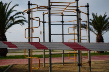 Bariyer bürokrasisi, spor sahasına giden bir geçidi kapatıyor spor malzemelerinin arka planında yakın plan çekiliyor. COVID-19 salgını. Palmiyeler, yeşil bitki örtüsü, sahil bulvarında bisiklet yolu..                               