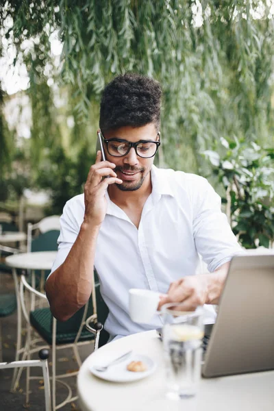 Olumlu ve çekici Afro Amerikalı işadamı bar üstünde laptop bazı işler yapıyor ve smartphone kullanarak kafede oturan
