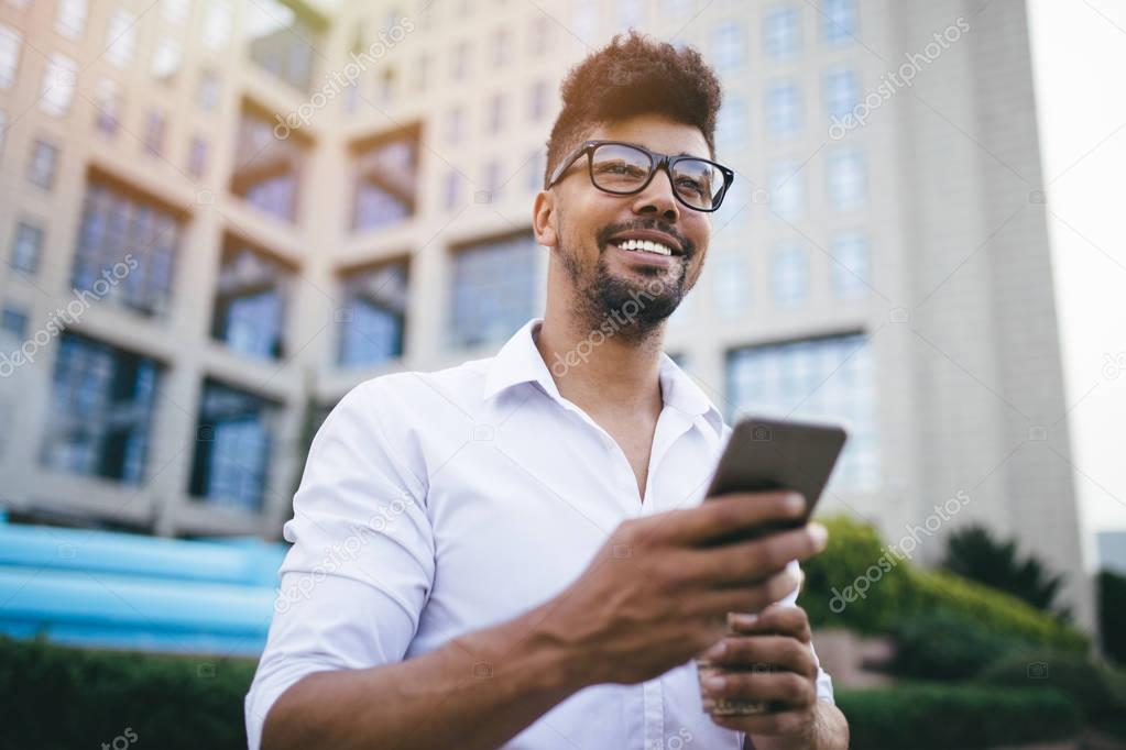 Young handsome Afro American man standing in front of huge modern business building smiling and using smartphone