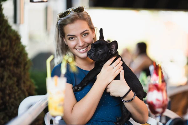 Beautiful young blonde woman with sunglasses enjoying with her french bulldog puppy in cafe bar