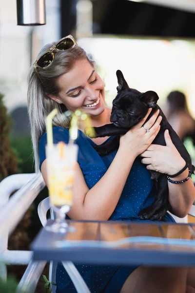 Beautiful young blonde woman with sunglasses enjoying with her french bulldog puppy in cafe bar