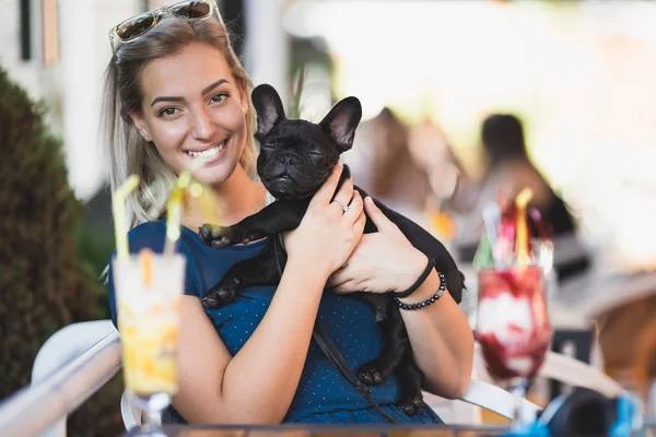 Beautiful young blonde woman with sunglasses enjoying with her french bulldog puppy in cafe bar