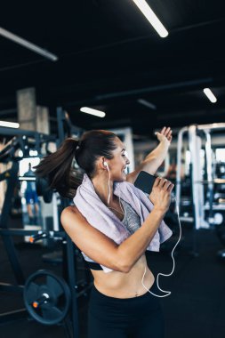 Genç ve çekici bir kadın modern spor salonunda başarılı bir idmandan sonra dans ediyor, gülümsüyor ve şarkı söylüyor. Telli kulaklıkları ve akıllı telefonuyla müzik dinliyor..