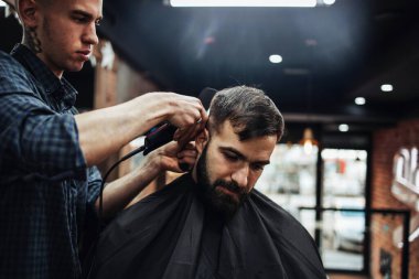 Yakışıklı bir genç adam saç ve sakal stili yaptırıyor ve profesyonel kuaför tarafından giydiriliyor..