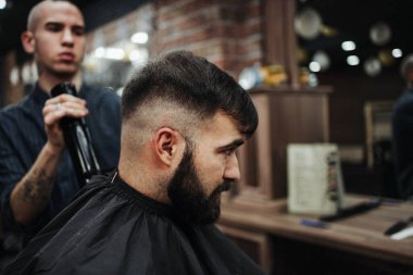 Yakışıklı bir genç adam saç ve sakal stili yaptırıyor ve profesyonel kuaför tarafından giydiriliyor..