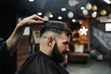 Yakışıklı bir genç adam saç ve sakal stili yaptırıyor ve profesyonel kuaför tarafından giydiriliyor..