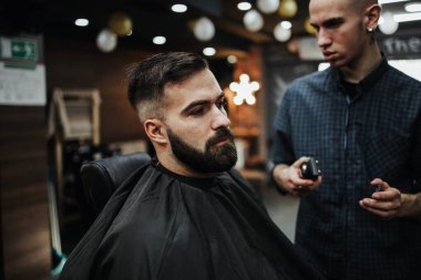 Yakışıklı bir genç adam saç ve sakal stili yaptırıyor ve profesyonel kuaför tarafından giydiriliyor..