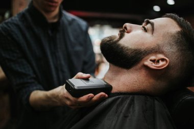 Yakışıklı bir genç adam saç ve sakal stili yaptırıyor ve profesyonel kuaför tarafından giydiriliyor..