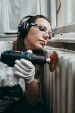 Güzel ve çalışkan orta yaşlı bir kadın eski evini ya da dairesini yenilerken kulaklıkla müzik dinliyor. Elinde profesyonel öğütücü ve daha sonra boyamak için radyatör tutuyor. Ev işi konseptini kendin yap..