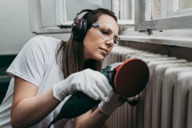 Güzel ve çalışkan orta yaşlı bir kadın eski evini ya da dairesini yenilerken kulaklıkla müzik dinliyor. Elinde profesyonel öğütücü ve daha sonra boyamak için radyatör tutuyor. Ev işi konseptini kendin yap..