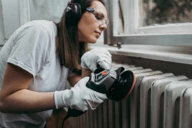 Güzel ve çalışkan orta yaşlı bir kadın eski evini ya da dairesini yenilerken kulaklıkla müzik dinliyor. Elinde profesyonel öğütücü ve daha sonra boyamak için radyatör tutuyor. Ev işi konseptini kendin yap..