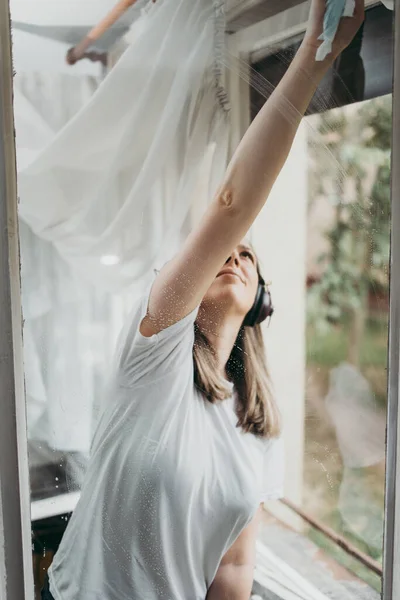Beautiful and diligent middle age handy woman cleaning windows at her home or apartment and listening to music with headphones. Do it yourself housework concept.