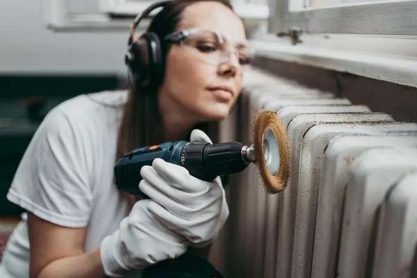 Beautiful and diligent middle age handy woman renovating her old home or apartment while listening to music with headphones. She is holding professional grinder and brushing radiator for later painting. Do it yourself housework concept.