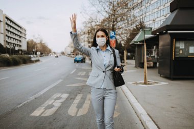 Koruyucu maskeli zarif bir iş kadını boş caddede tek başına duruyor ve kaldırılmış elleriyle otobüs ya da taksi çağırıyor. Corona virüsü veya Covid-19 yaşam tarzı konsepti.