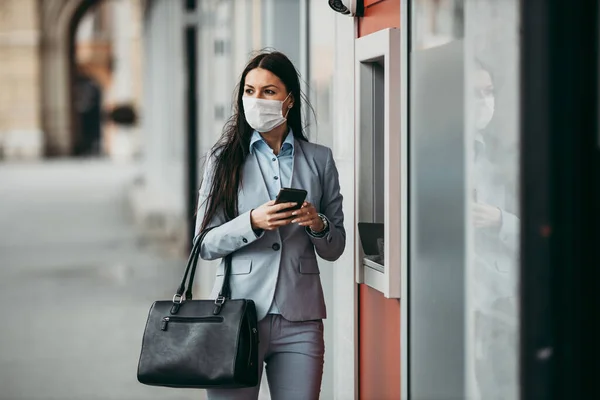 Elegant business woman with protective mask standing on city street and using ATM machine to withdraw cash. Corona or Covid-19 virus pandemic concept.