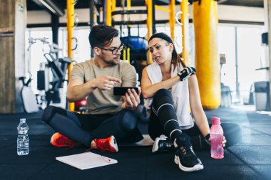 Erkek fitness eğitmeni, akıllı telefonuyla spor yaparken yerde genç ve çekici bir kadınla oturup dinleniyor..