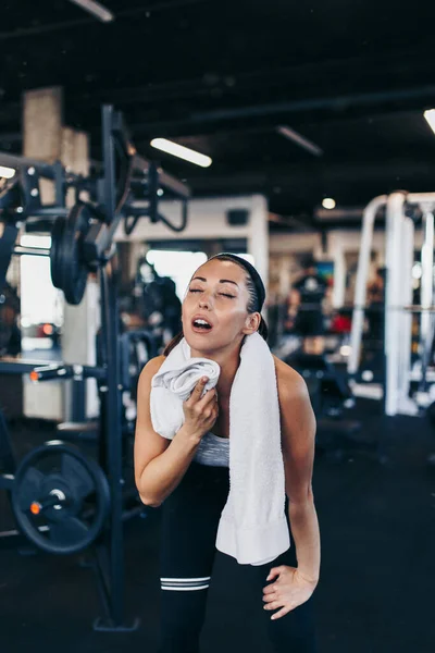 Young exhausted woman relaxing in fitness gym after successful workout wipes sweat from the forehead with white towel.