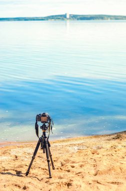 Bir kamera bir tripod üzerinde kum sahil deniz manzarası kaldırır. Manzara çekim sürecinde fotoğrafik donanımlar. Kamera fotoğraf çekerken veya hızlandırılmış