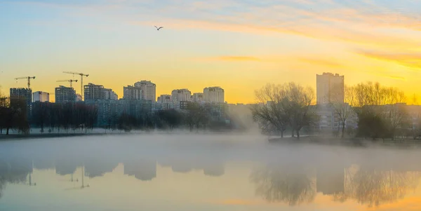 Heavy fog on water in morning. City skyline with multistory buildings on river bank