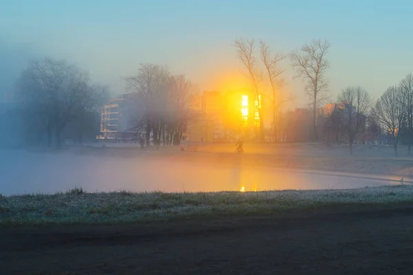 Alone silhouette of a cyclist riding in a strong fog along the river bank in a city park early in the morning. Rays of the sun in the reflection of the windows of buildings cut through a thick fog