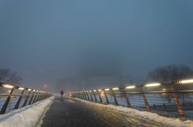 Güçlü bir sisin içinde karanlıkta bir yaya köprüsü buradan geçen bir adam silüeti. Küpeşte entegre lambalar ile yolu onun için aydınlatmak