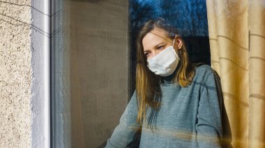 Gazlı bez maskeli bir kadın pencereden dışarı bakıyor. Anti-virüs ev izolasyonu konsepti. Salgın sırasında sağlığınızı koruyun..