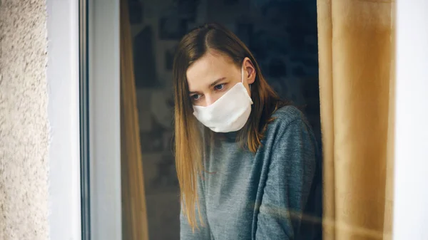A woman in a gauze mask looks through the window. Anti-virus home isolation concept. Protect your health during the epidemic.