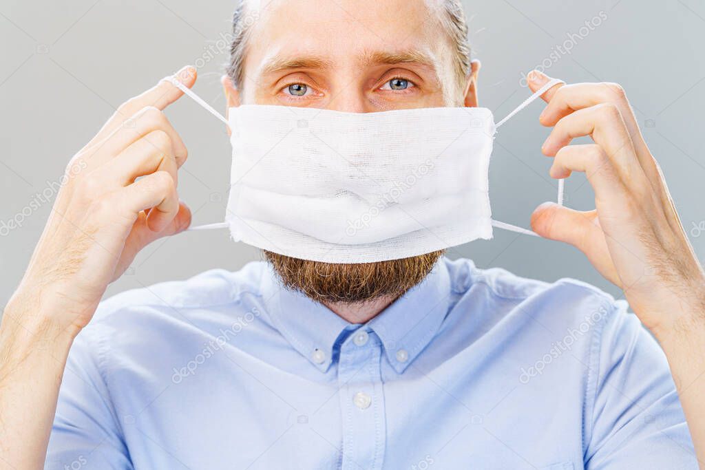 White bearded man in blue shirt puts on a mask from air pollution and coronavirus Covid-19, looking at camera. Studio shot on gray background