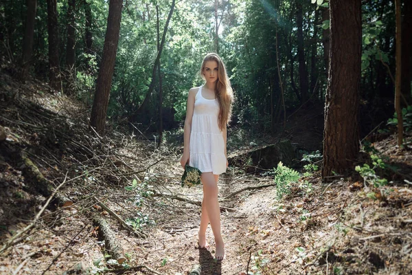 Menina com um buquê de flores e um vestido branco em uma floresta ensolarada — Fotografia de Stock