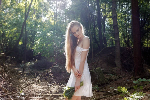 Menina com um buquê de flores e um vestido branco em uma floresta ensolarada — Fotografia de Stock