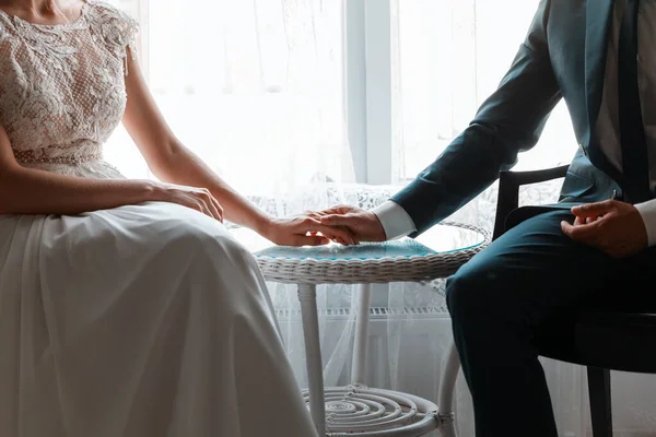 couple in wedding dresses sitting at the table and holding hands against the background of the window, without a face