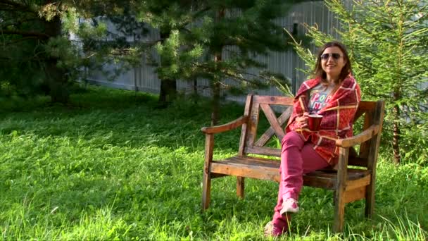 Chica sonriente envuelta en una cálida taza de té a cuadros, sentada en el banco del parque — Vídeos de Stock