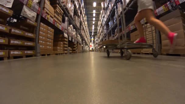 Funny Young Girl en mini falda monta carro de la compra, ir a la distancia en la tienda de muebles almacén entre filas de estantes con cajas de cartón — Vídeos de Stock