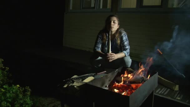 Chica sentada junto al fuego en la noche, agregando leña al fuego — Vídeos de Stock