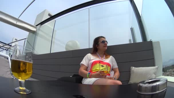 Bella ragazza in occhiali da sole, minigonna e t-shirt situata sul divano della terrazza del balcone dell'hotel e cocktail, la ragazza gode della vista sul paesaggio — Video Stock