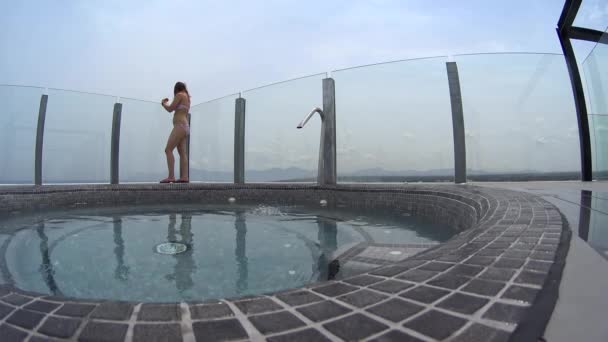 Bella ragazza in occhiali da sole e bikini rosa stare sul bordo della jacuzzi sul tetto dell'hotel e bere cocktail, accendere la vasca idromassaggio, sedersi e mettere le gambe in acqua — Video Stock