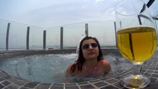 Dziewczyna w okulary relaksującą kąpiel w wannie z hydromasażem, pić piwo koktajl na skraju jacuzzi na dachu hotelu, piękny widok na krajobraz — Wideo stockowe