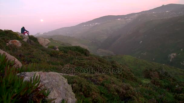 Дівчина п'є пляшку, сидячи на гірській скелі, спостерігаючи за вечірнім місяцем — стокове відео