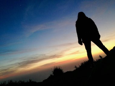 Okyanus su üzerinde gün batımını izlemek kızla dağ üst uçurum büyüdü, el ile bir sırt çantası siluet duran