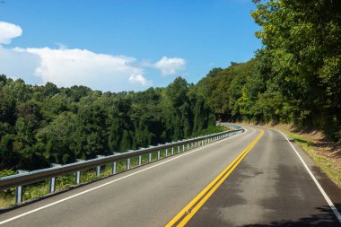 Asphalt road along the trees.  Beautiful empty highway with yellow lines between green trees on a summer warm day with blue sky and fluffy clouds