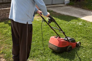 Bir işçi elektrikli çim biçme makinesiyle yeşil bir çim biçiyor. Çim biçme makinesi kırmızı. Yatay çerçeve
