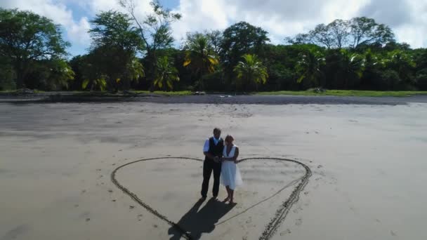 Zdjęcie lotnicze pary sukni ślubnych na tropikalnej plaży Playa Arenillas w Kostaryce z sercem wciągniętym w piasek — Wideo stockowe